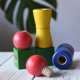 Nic screws and nuts colorful wooden toy threaded blocks and shapes arranged on a white wooden surface, including red spherical pieces, a green block, a yellow cone-shaped piece, and a blue cylindrical piece, with a blurred green leaf in the background.