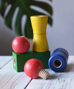 Nic screws and nuts colorful wooden toy threaded blocks and shapes arranged on a white wooden surface, including red spherical pieces, a green block, a yellow cone-shaped piece, and a blue cylindrical piece, with a blurred green leaf in the background.