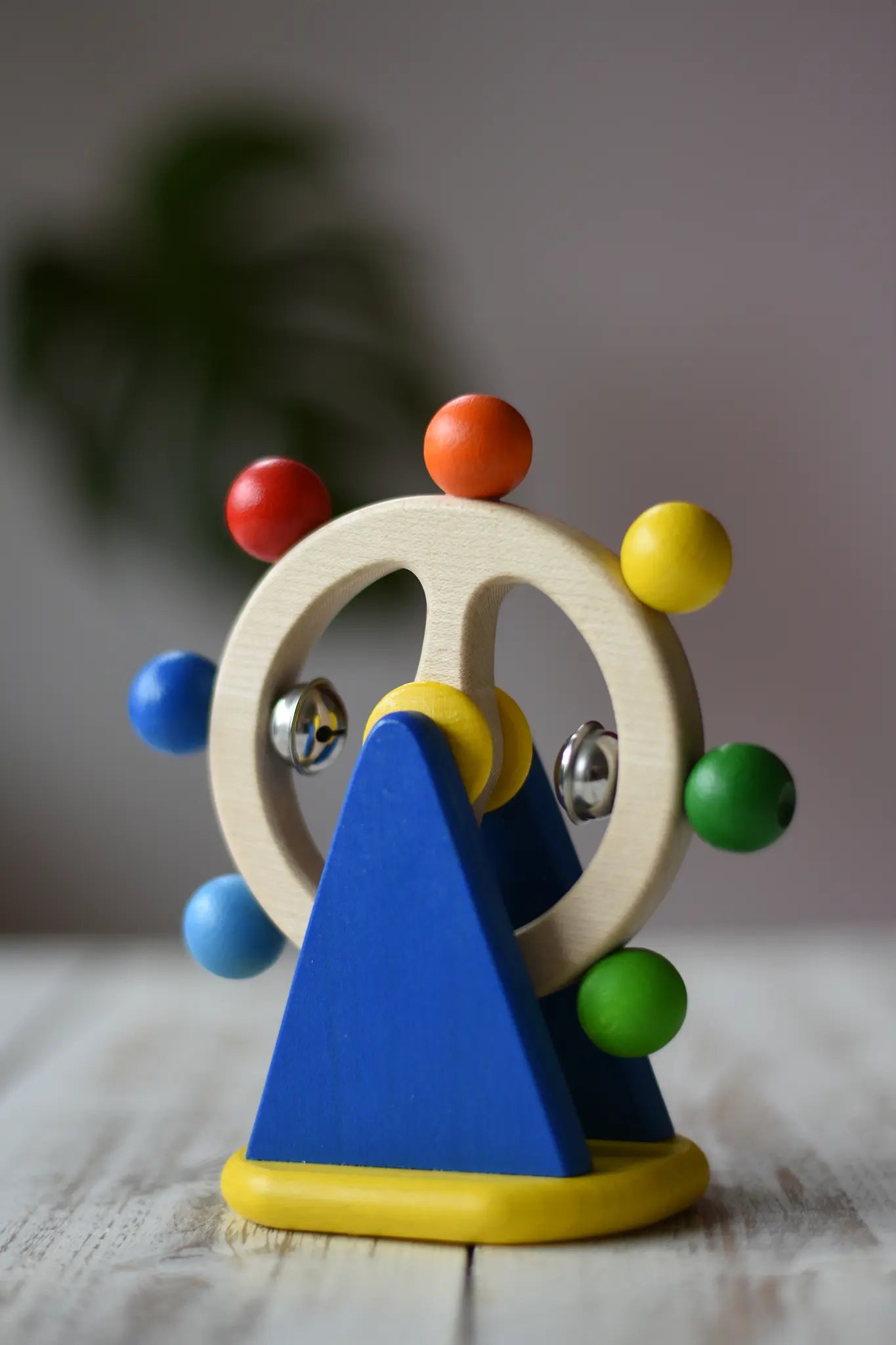 Nic Toys ferris wheel colorful wooden toy for infants and toddlers, featuring a natural wood wheel with colorful painted balls, two silver metal balls, and a blue triangular support on a yellow base, set on a light wooden tabletop.