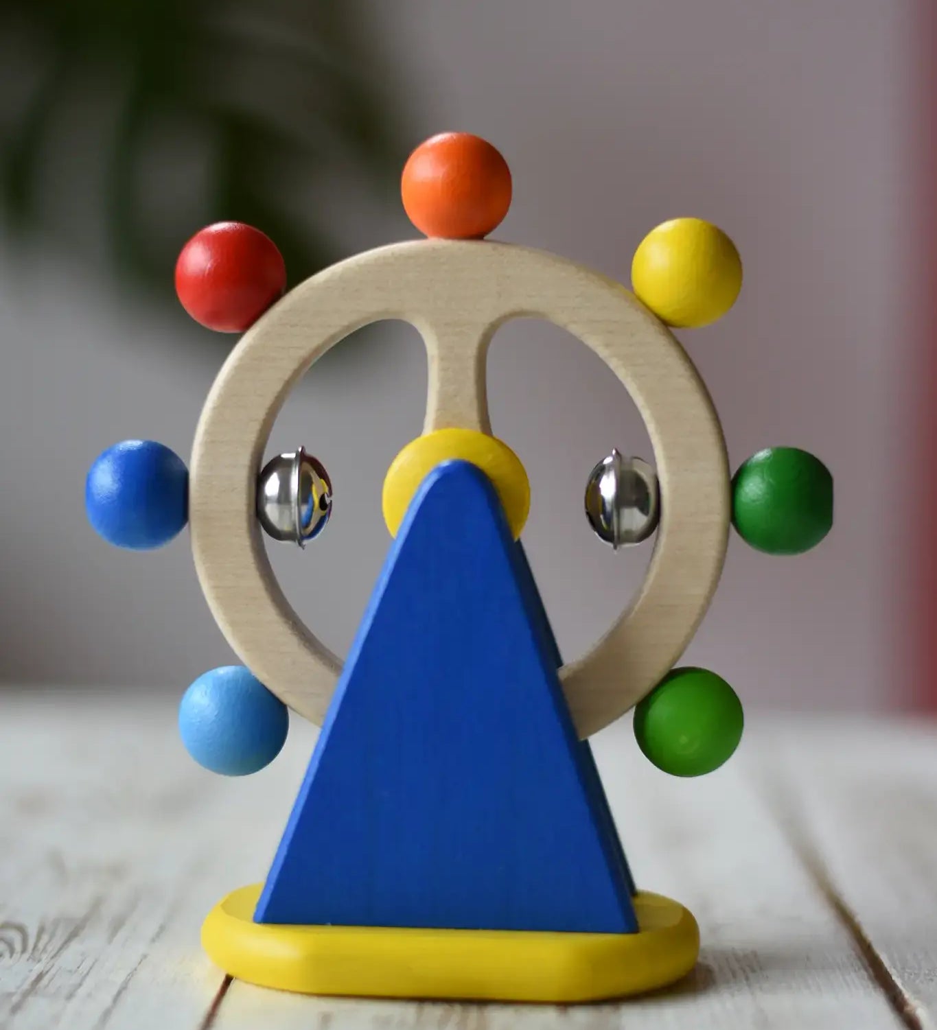 Nic Toys ferris wheel colorful wooden toy for infants and toddlers, featuring a natural wood wheel with colorful painted balls, two silver metal balls, and a blue triangular support on a yellow base, set on a light wooden tabletop.