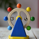 Nic Toys ferris wheel colorful wooden toy for infants and toddlers, featuring a natural wood wheel with colorful painted balls, two silver metal balls, and a blue triangular support on a yellow base, set on a light wooden tabletop.