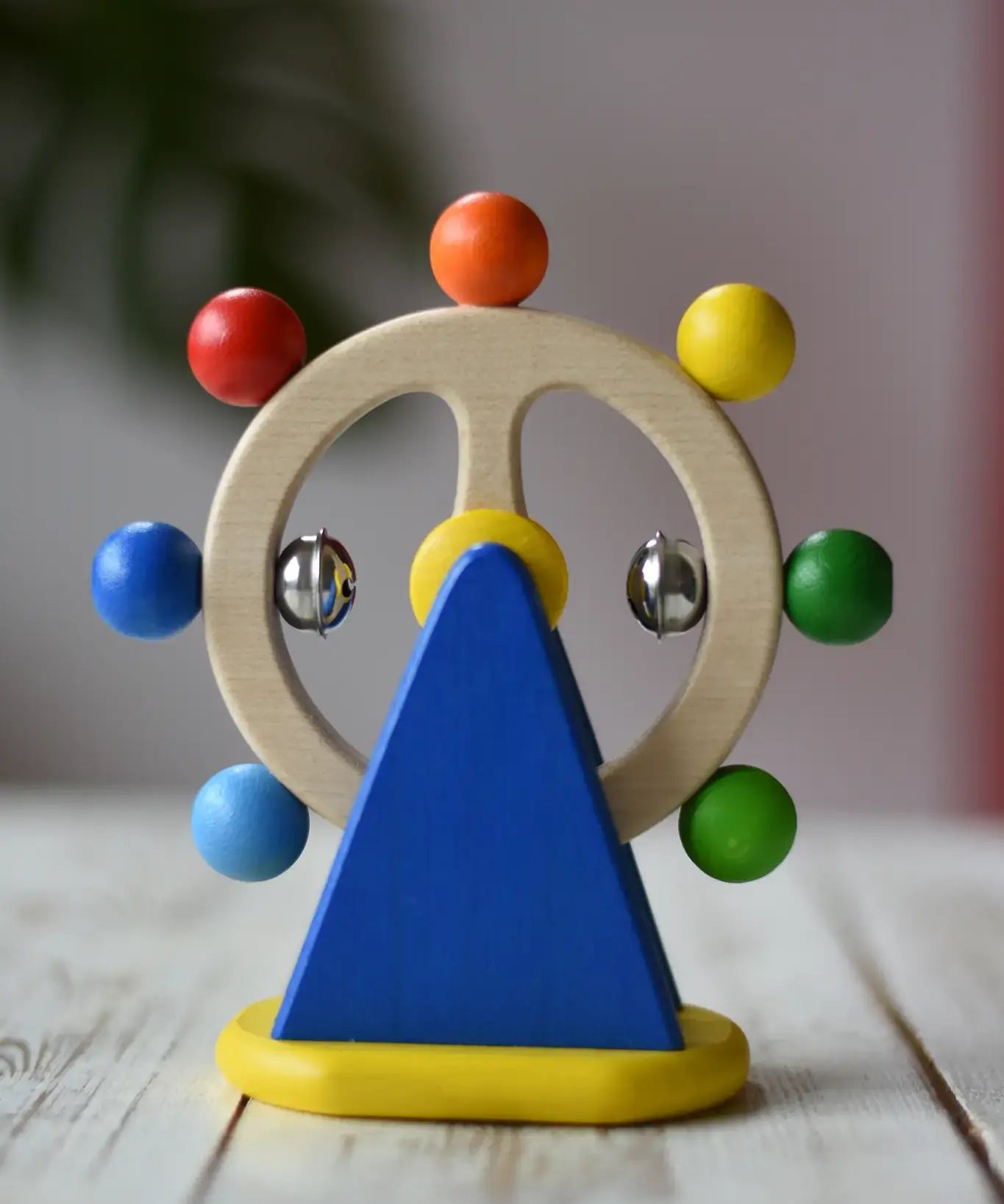 Nic Toys ferris wheel colorful wooden toy for infants and toddlers, featuring a natural wood wheel with colorful painted balls, two silver metal balls, and a blue triangular support on a yellow base, set on a light wooden tabletop.