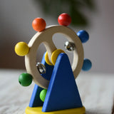 Nic Toys ferris wheel colorful wooden toy for infants and toddlers, featuring a natural wood wheel with colorful painted balls, two silver metal balls, and a blue triangular support on a yellow base, set on a light wooden tabletop.