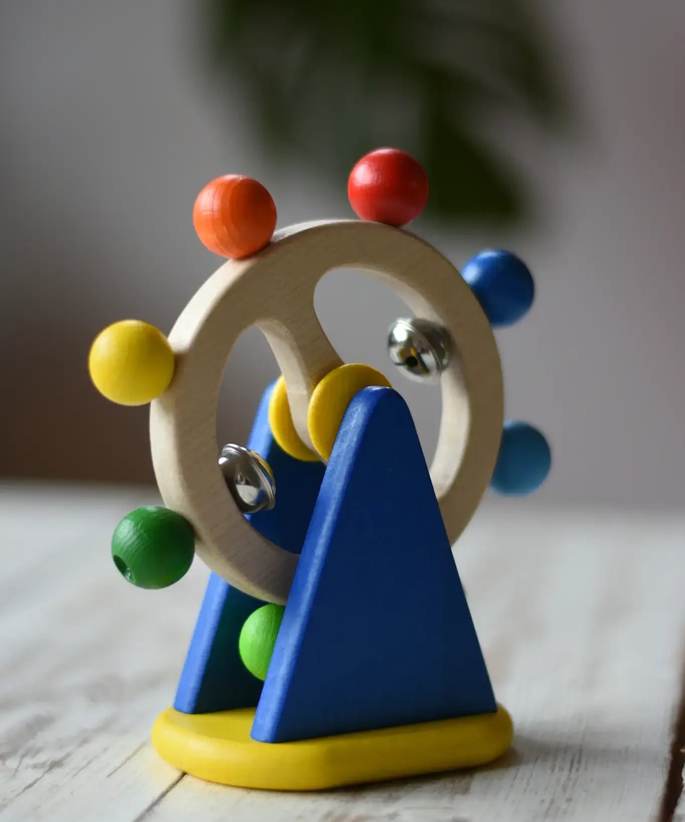 Nic Toys ferris wheel colorful wooden toy for infants and toddlers, featuring a natural wood wheel with colorful painted balls, two silver metal balls, and a blue triangular support on a yellow base, set on a light wooden tabletop.
