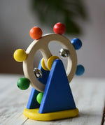 Nic Toys ferris wheel colorful wooden toy for infants and toddlers, featuring a natural wood wheel with colorful painted balls, two silver metal balls, and a blue triangular support on a yellow base, set on a light wooden tabletop.
