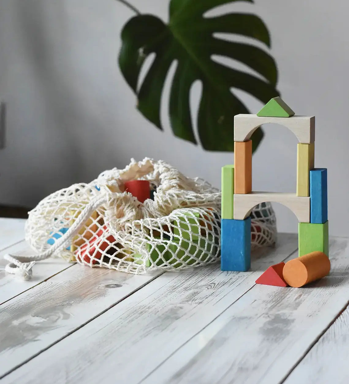 White mesh drawstring bag filled with Glückskäfer colorful wooden blocks next to a tower of wooden blocks. A large green monstera leaf is visible in the soft-focus background. Glückskäfer Wooden Bio Geometric Building Blocks - 58 Piece Se.
