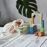White mesh drawstring bag filled with Glückskäfer colorful wooden blocks next to a tower of wooden blocks. A large green monstera leaf is visible in the soft-focus background. Glückskäfer Wooden Bio Geometric Building Blocks - 58 Piece Se.