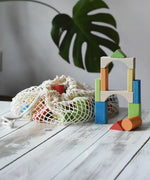 White mesh drawstring bag filled with Glückskäfer colorful wooden blocks next to a tower of wooden blocks. A large green monstera leaf is visible in the soft-focus background. Glückskäfer Wooden Bio Geometric Building Blocks - 58 Piece Se.