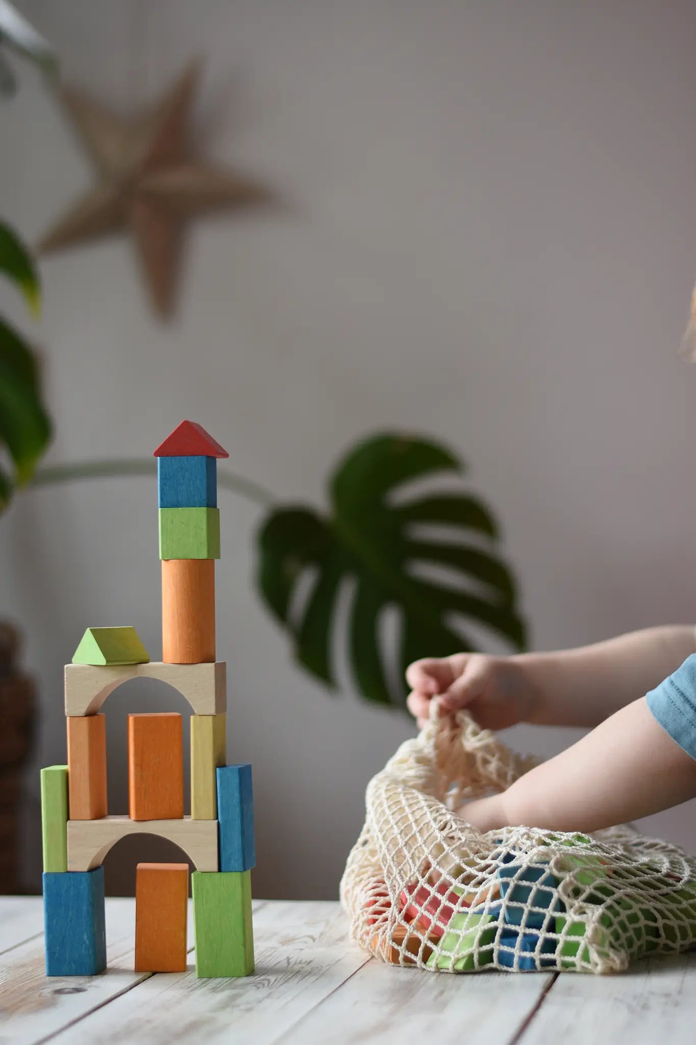 Child taking Glückskäfer Wooden Bio Geometric Building Blocks out of the net storage bag next to a tower created with the blocks.  
