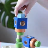 Child's hand placing a wooden straw into a block from the Nic set of colorful wooden screws and blocks arranged wooden tabletop, with a blurred green plant leaf in the background.