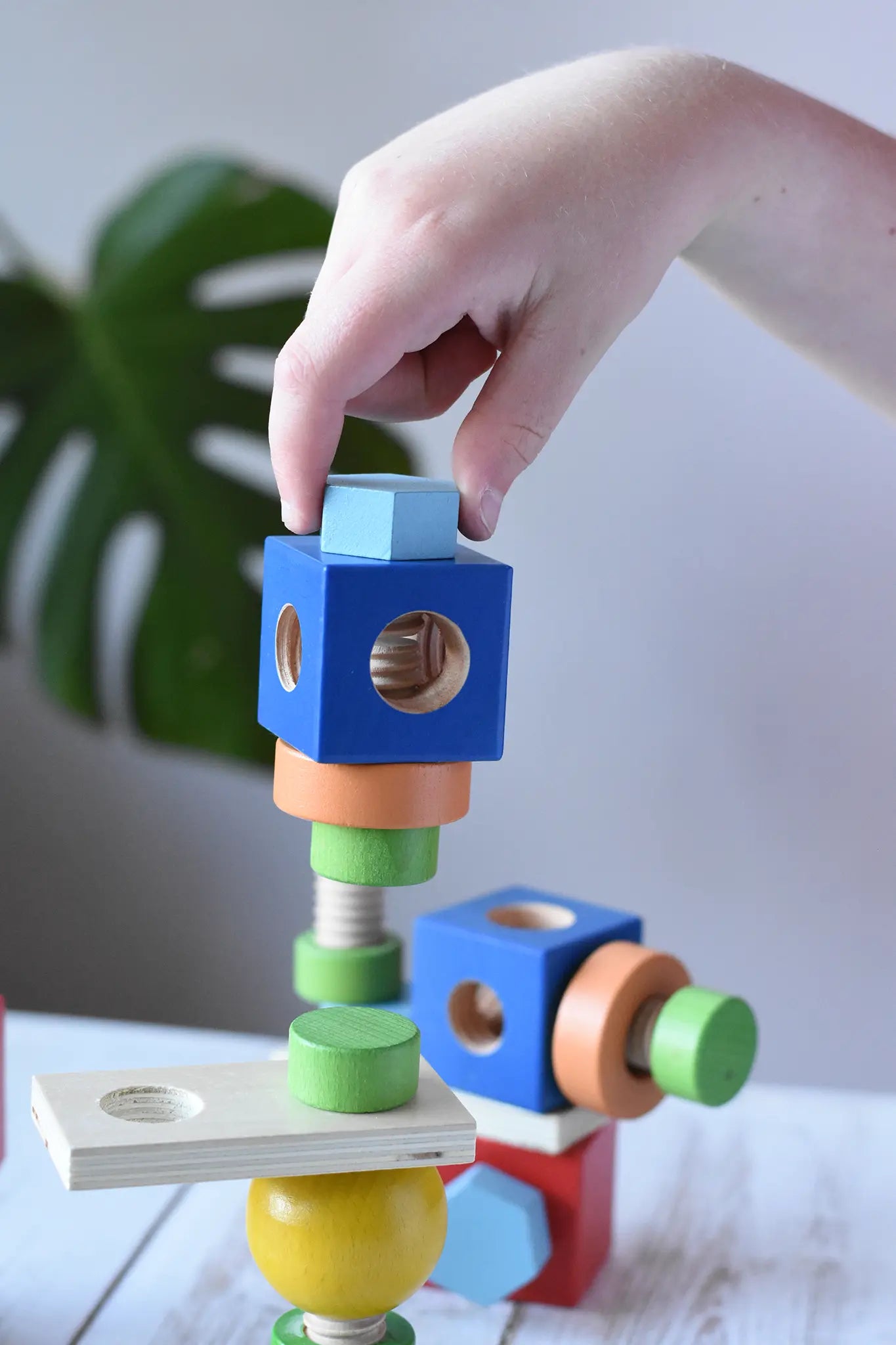 Child's hand placing a wooden straw into a block from the Nic set of colorful wooden screws and blocks arranged wooden tabletop, with a blurred green plant leaf in the background.