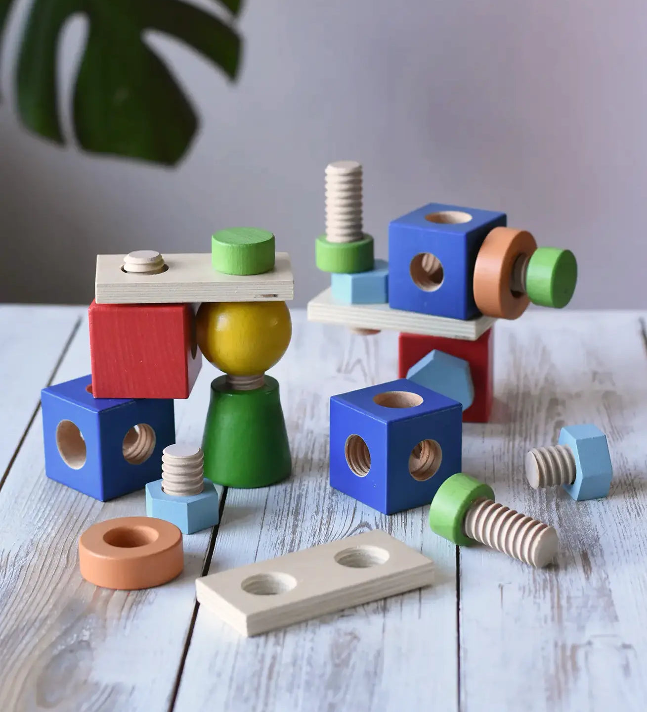 Nic set of colorful wooden screws and blocks arranged wooden tabletop, with a blurred green plant leaf in the background.