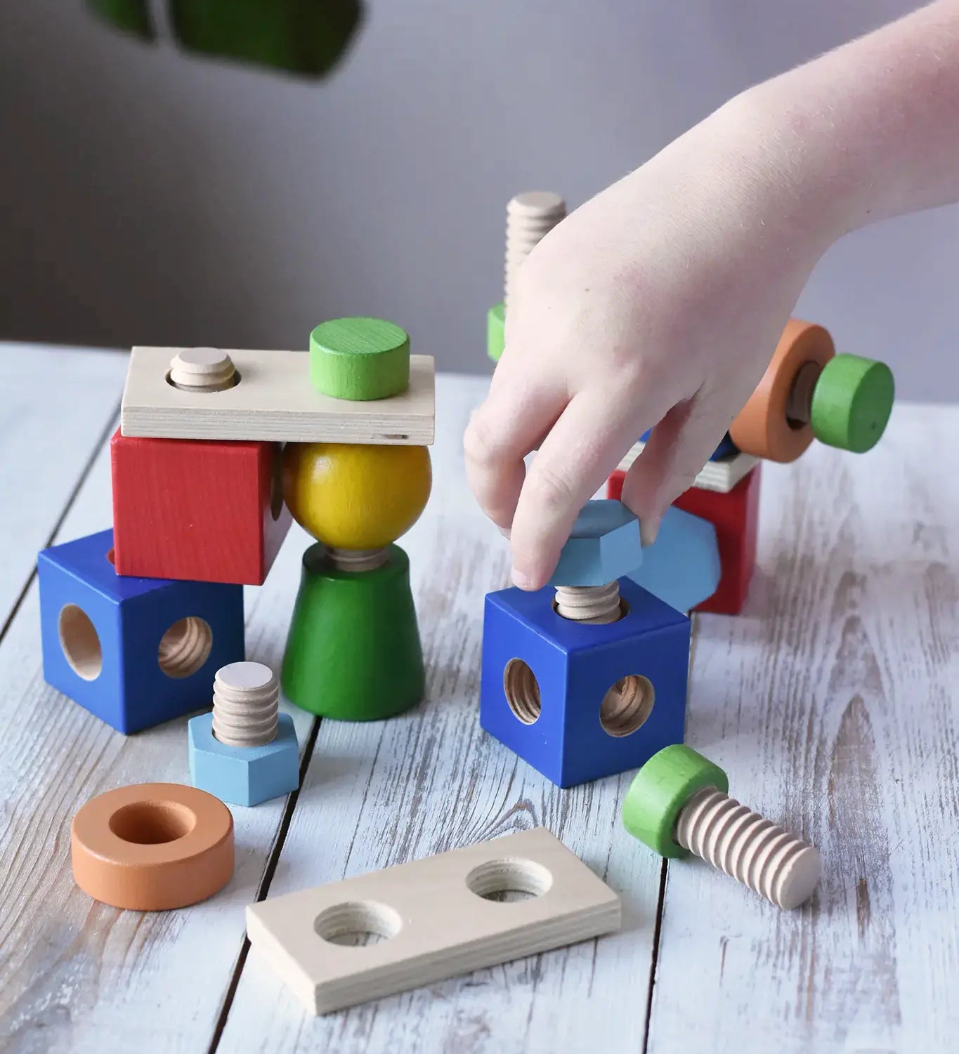 A child's hand is playing with a set of Nic set of colorful wooden screws and blocks arranged wooden tabletop, with a blurred green plant leaf in the background. The set includes blue, red, green, yellow, orange, and natural wood pieces.