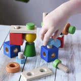 A child's hand is playing with a set of Nic set of colorful wooden screws and blocks arranged wooden tabletop, with a blurred green plant leaf in the background. The set includes blue, red, green, yellow, orange, and natural wood pieces.
