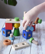 A child's hand is playing with a set of Nic set of colorful wooden screws and blocks arranged wooden tabletop, with a blurred green plant leaf in the background. The set includes blue, red, green, yellow, orange, and natural wood pieces.