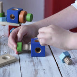 Close-up of a child's hands assembling colorful wooden blocks and threaded screws on a light rustic table. The toy set includes blue blocks, green pegs, orange rings, and natural wood pieces.