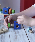 Close-up of a child's hands assembling colorful wooden blocks and threaded screws on a light rustic table. The toy set includes blue blocks, green pegs, orange rings, and natural wood pieces.