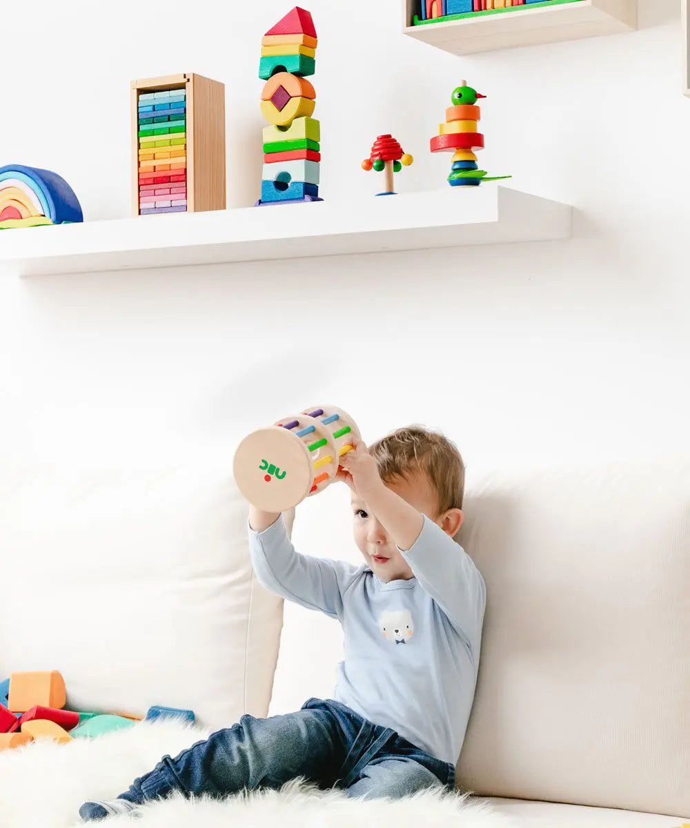 Nic Toys Rattle n Roll - a wooden musical rolling wheel toy for babies, shown with its natural finish, rainbow rungs and wooden marbles in a playroom setting.