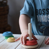 Child playing with a Nic wooden peg-hole stacking tower toy on a white wooden table. The toy features a natural wood peg and rings in red, yellow, orange, green, and blue.