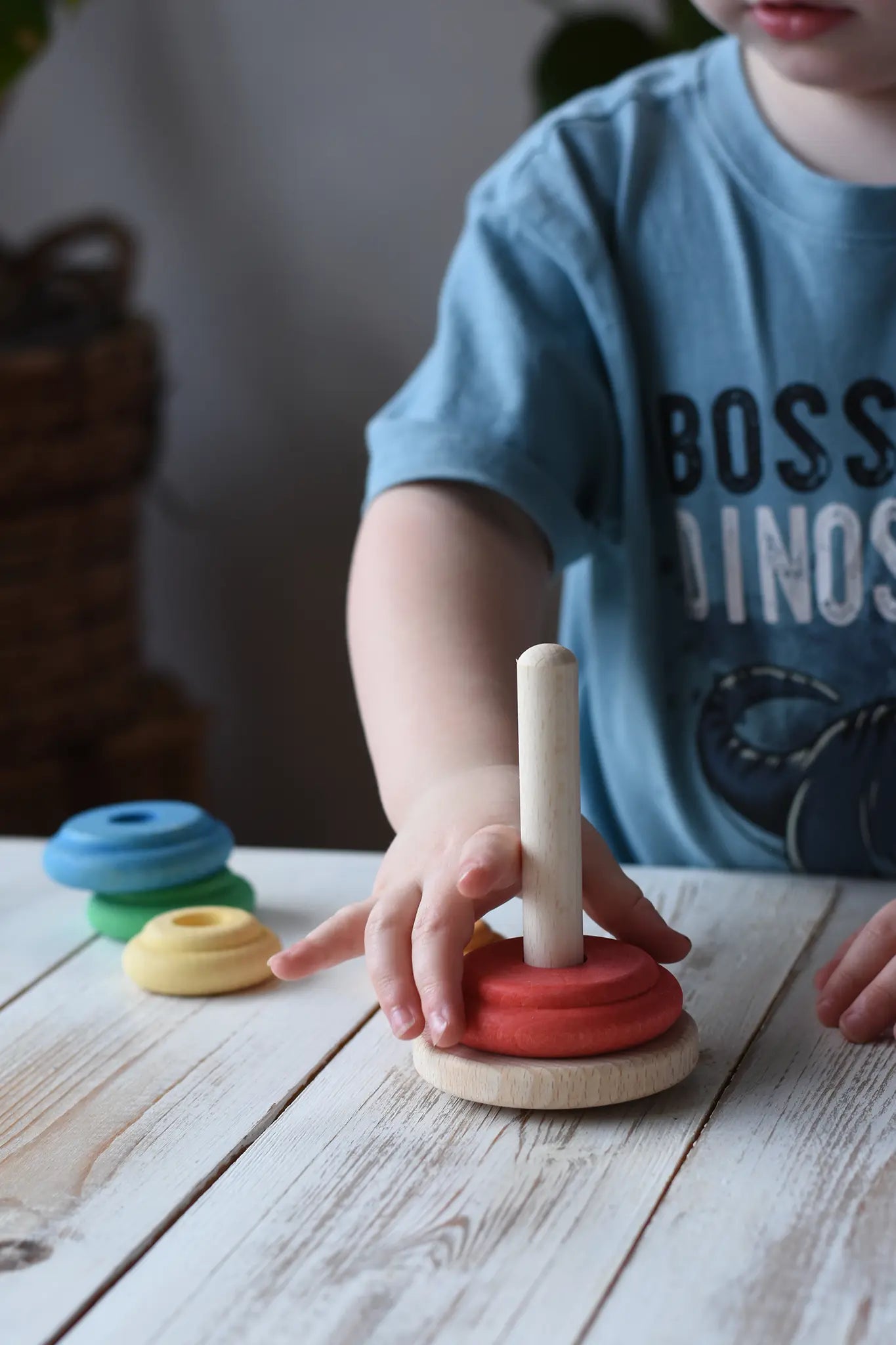 Child playing with a Nic wooden peg-hole stacking tower toy on a white wooden table. The toy features a natural wood peg and rings in red, yellow, orange, green, and blue.