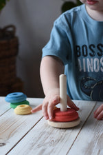 Child playing with a Nic wooden peg-hole stacking tower toy on a white wooden table. The toy features a natural wood peg and rings in red, yellow, orange, green, and blue.