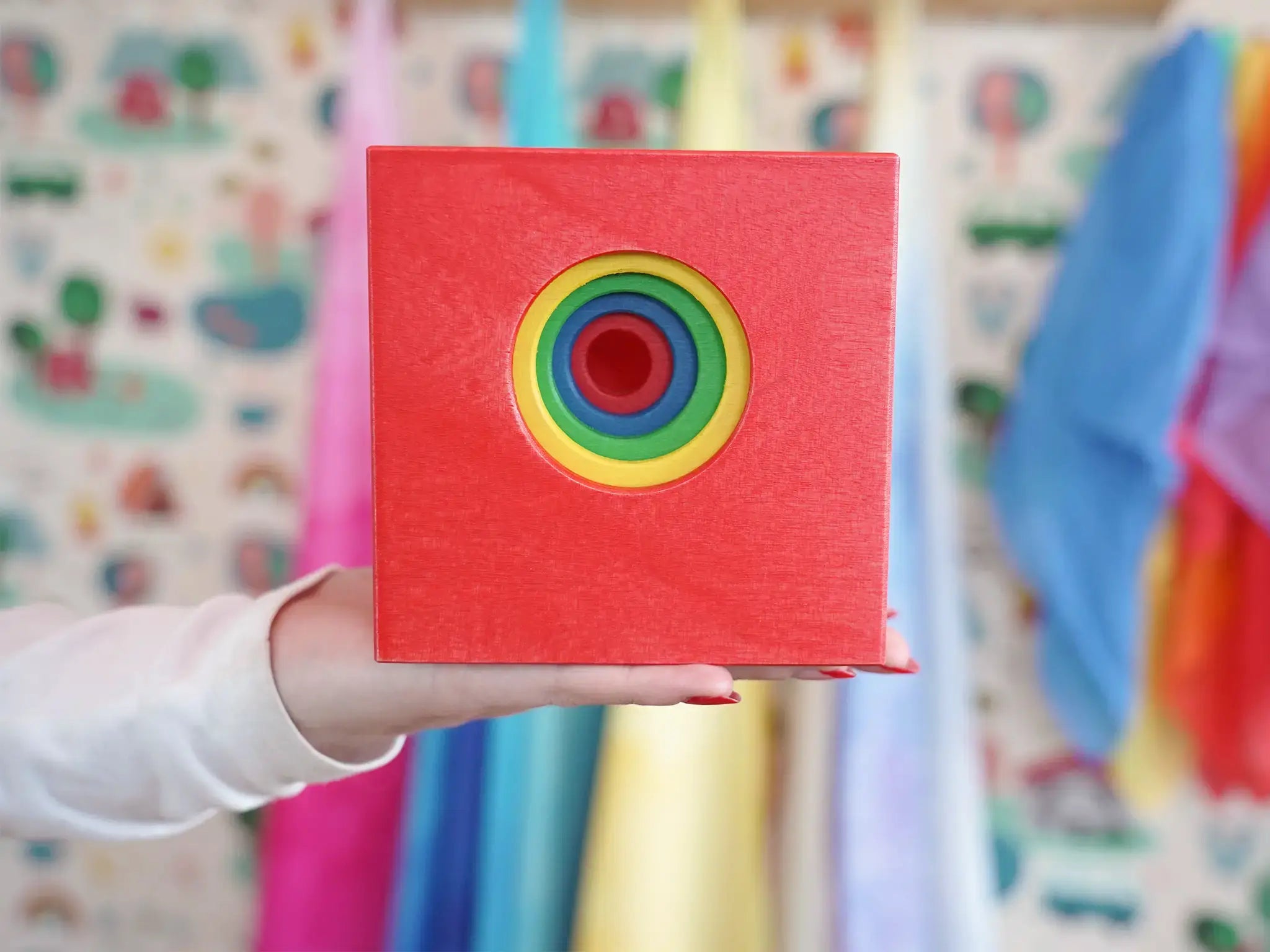 A hand holding a red wooden toy box with smaller boxes nested inside with a hole in each one making it look like a colorful rainbow concentric circle target on the front, set against a softly blurred background of children's play materials.
