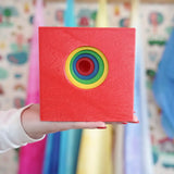 A hand holding a red wooden toy box with smaller boxes nested inside with a hole in each one making it look like a colorful rainbow concentric circle target on the front, set against a softly blurred background of children's play materials.