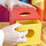 A close-up of a person's hands interacting with a colorful wooden Montessori-style box with a red top and a yellow drawer, set against a soft, blurred background of bright colors.
