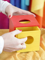 A close-up of a person's hands interacting with a colorful wooden Montessori-style box with a red top and a yellow drawer, set against a soft, blurred background of bright colors.