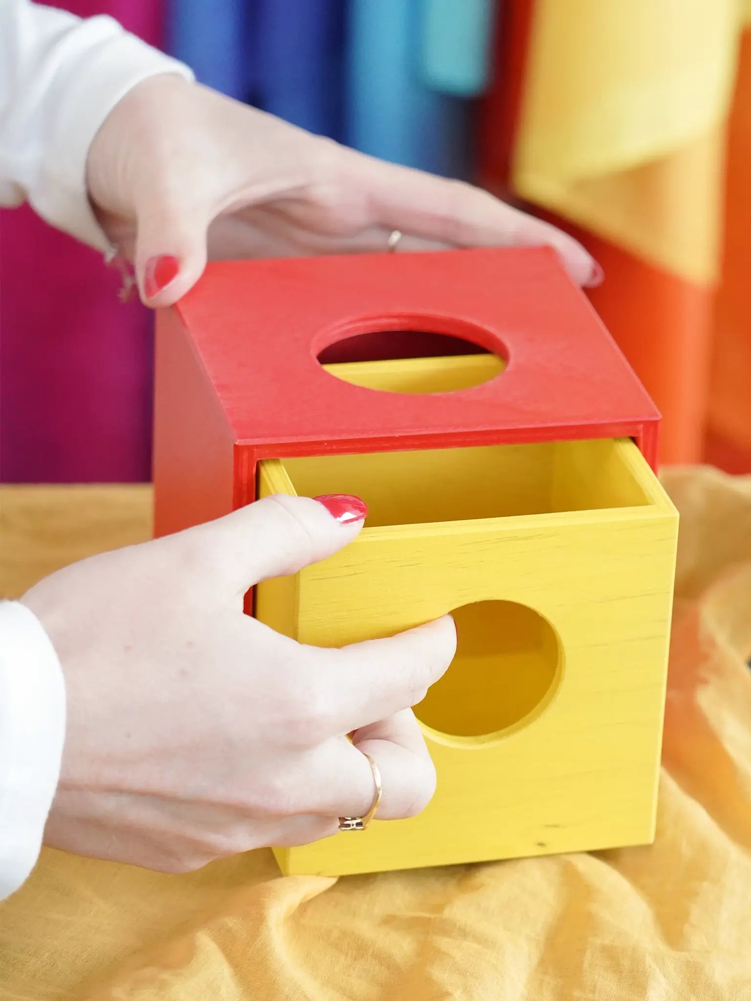 A close-up of a person's hands interacting with a colorful wooden Montessori-style box with a red top and a yellow drawer, set against a soft, blurred background of bright colors.