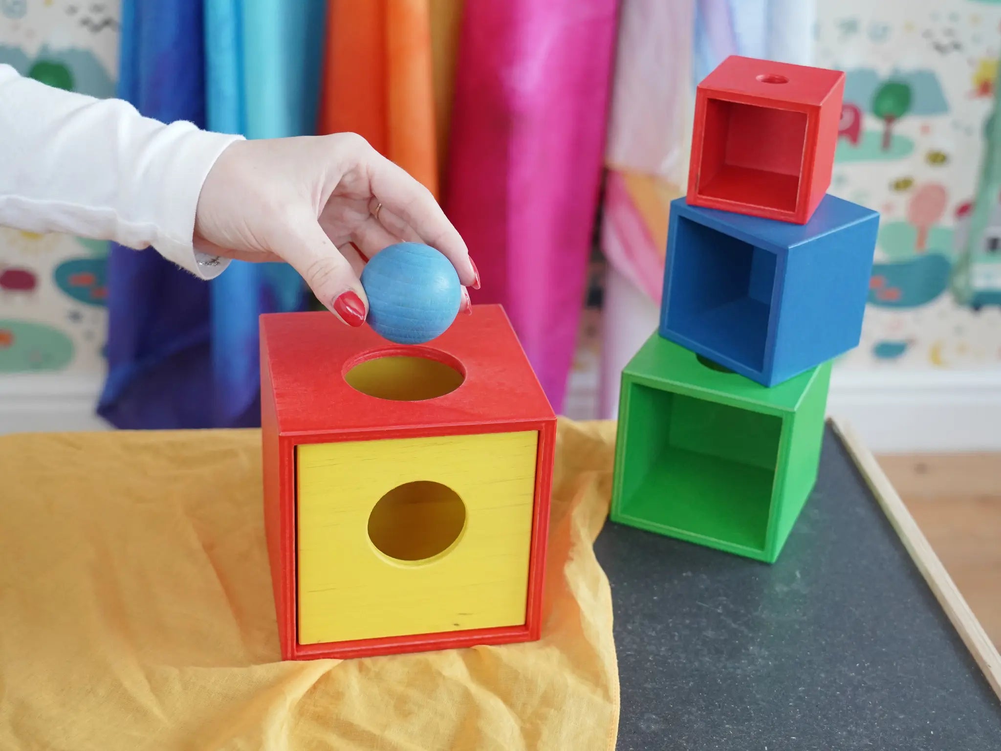 Nic Toys Stacking boxes set. A hand  places a blue ball into the  red and yellow wooden box put togethr like a  shape-sorting box. 