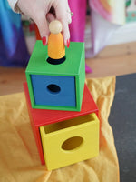 A colorful Nic Toys wooden stacking and nesting box toy with a being used as a permanence box to insert a small wooden peg doll.