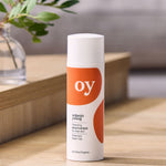 The Oy organic cleaning moisturiser for clear skin with prebiotics and green tea, is stood on a brown table with a plant in the background