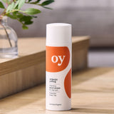 The Oy organic cleaning moisturiser for clear skin with prebiotics and green tea, is stood on a brown table with a plant in the background