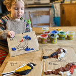 Child showing art he has created using Okonorm play dough. The child has recreated different birds on cardboard by using the different coloured play doughs. The child is sat on a chair at the kitchen table. He has one piece of art in his hands, the other three are on the table.