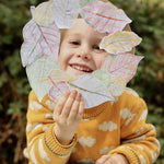 Child holding a paper leaf wreath he has made with leaf rubbings, having used Okonorm wax blocks. The child is holding the wreath in front of his face, you can see the child's face in the hole of the wreath. The child is wearing a yellow knitted jumper with a white cloud pattern and is sat outside.