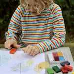 Okonorm wax blocks being used by a child. The child is sat outside in the garden and is creating leaf rubbings with the wax blocks. The child is wearing a striped hoodie and has curly, blonde, above shoulder length hair.