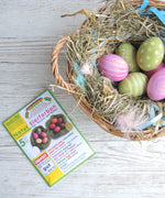ÖkoNORM nawaro natural egg dye set placed on a light wooden surface beside a wicker basket filled with hay and dyed Easter eggs. The packaging is green and white, featuring an image of naturally dyed eggs in shades of red, pink, yellow, and brown. The label highlights that the dyes are made from natural ingredients. The basket contains decorated eggs in pastel shades of pink and green, some with polka dot and striped patterns, nestled among hay, feathers, and blue ribbon