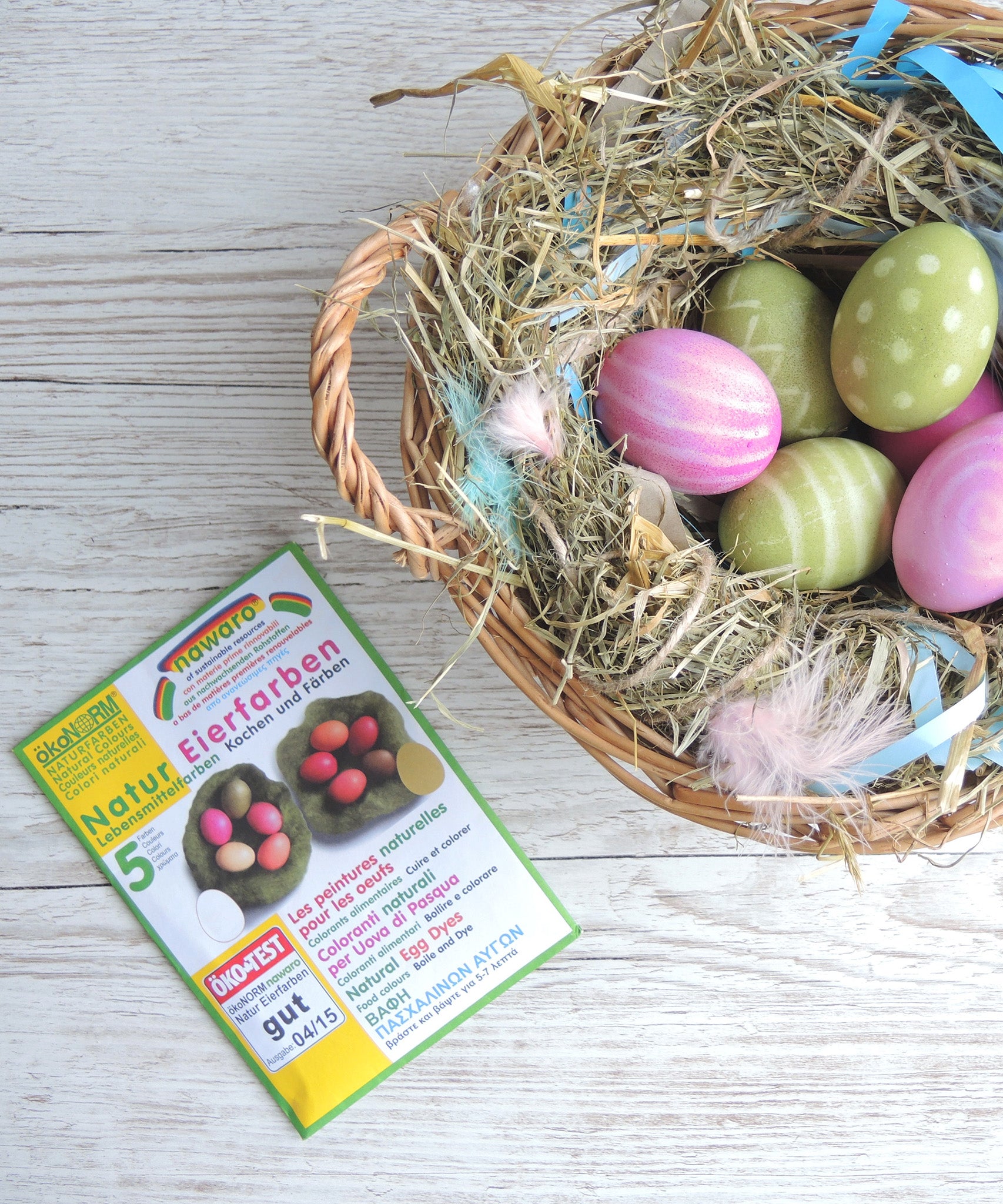 ÖkoNORM nawaro natural egg dye set placed on a light wooden surface beside a wicker basket filled with hay and dyed Easter eggs. The packaging is green and white, featuring an image of naturally dyed eggs in shades of red, pink, yellow, and brown. The label highlights that the dyes are made from natural ingredients. The basket contains decorated eggs in pastel shades of pink and green, some with polka dot and striped patterns, nestled among hay, feathers, and blue ribbon