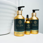 The Olew Curl Defining Gel, Original Oil and Curl Cream in gold and dark green bottles, stood on a white table with a white large bowl next to the bottles.