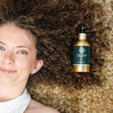 A person with brown curly hair laying down on a light coloured curly rug, with their hair on the rug and the Olew Hold Spray laid on top of their curls.