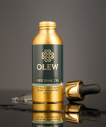 The Gold and black pipette top is laid behind the Olew Original Hair Oil bottle, on a dark grey background