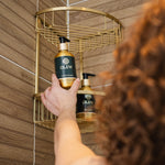 A person reaching for the Olew conditioner and shampoo bottles which are placed in a gold coloured shower caddy