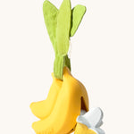 The Ana Banana Pacifier Holder on a cream background. The image shows an Oli & Carol banana teething toy half way inside the holder. The banana is smooth and has a green muslin leaf attachment on the top to tie the holder to a pram handle, high chair etc.