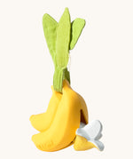 The Ana Banana Pacifier Holder on a cream background. The image shows an Oli & Carol banana teething toy half way inside the holder. The banana is smooth and has a green muslin leaf attachment on the top to tie the holder to a pram handle, high chair etc.