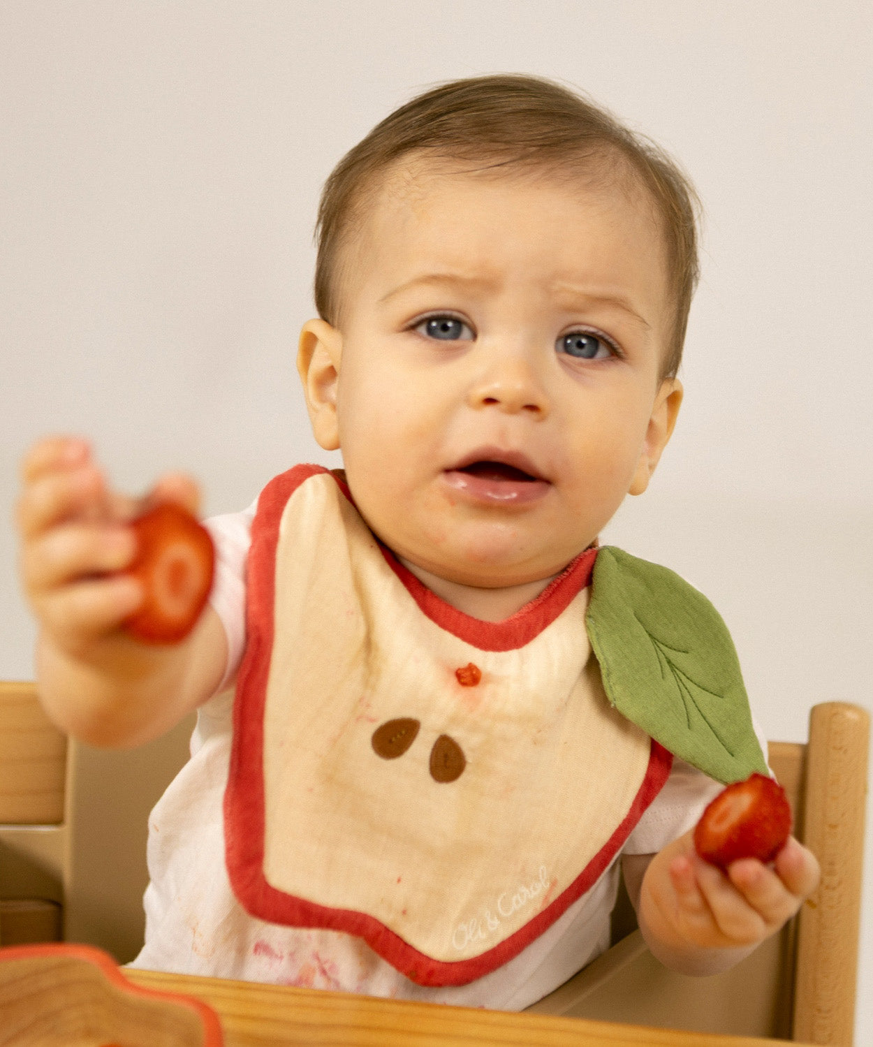 A baby sitting in a wooden high chair eating strawberries and wearing the Oli & Carol Peppa the apple baby bib available at Babipur. 