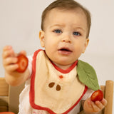A baby sitting in a wooden high chair eating strawberries and wearing the Oli & Carol Peppa the apple baby bib available at Babipur. 