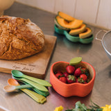 Oli & Carol Peppa the apple wooden bowl and spoon set available at Babipur.  The bowl is in a shape of a apple cut in half and features a decorative green leaf on the bowl. Bowl is on a kitchen surface filled with fruit