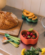 Oli & Carol Peppa the apple wooden bowl and spoon set available at Babipur.  The bowl is in a shape of a apple cut in half and features a decorative green leaf on the bowl. Bowl is on a kitchen surface filled with fruit