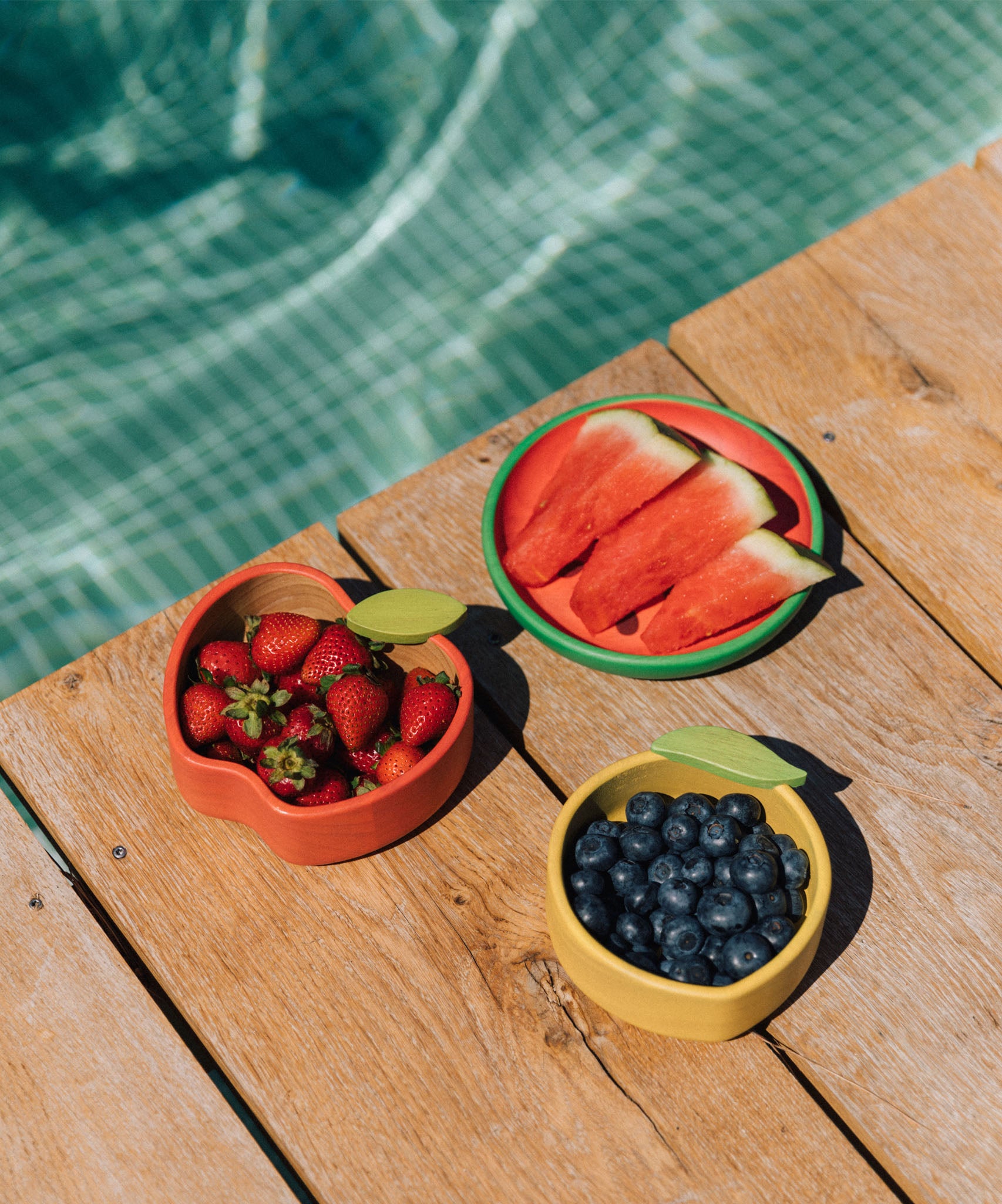 The Oli & Carol Peppa the apple wooden bowl next to a lemon bowl and watermelon plate filled with fruit. available at Babipur. 